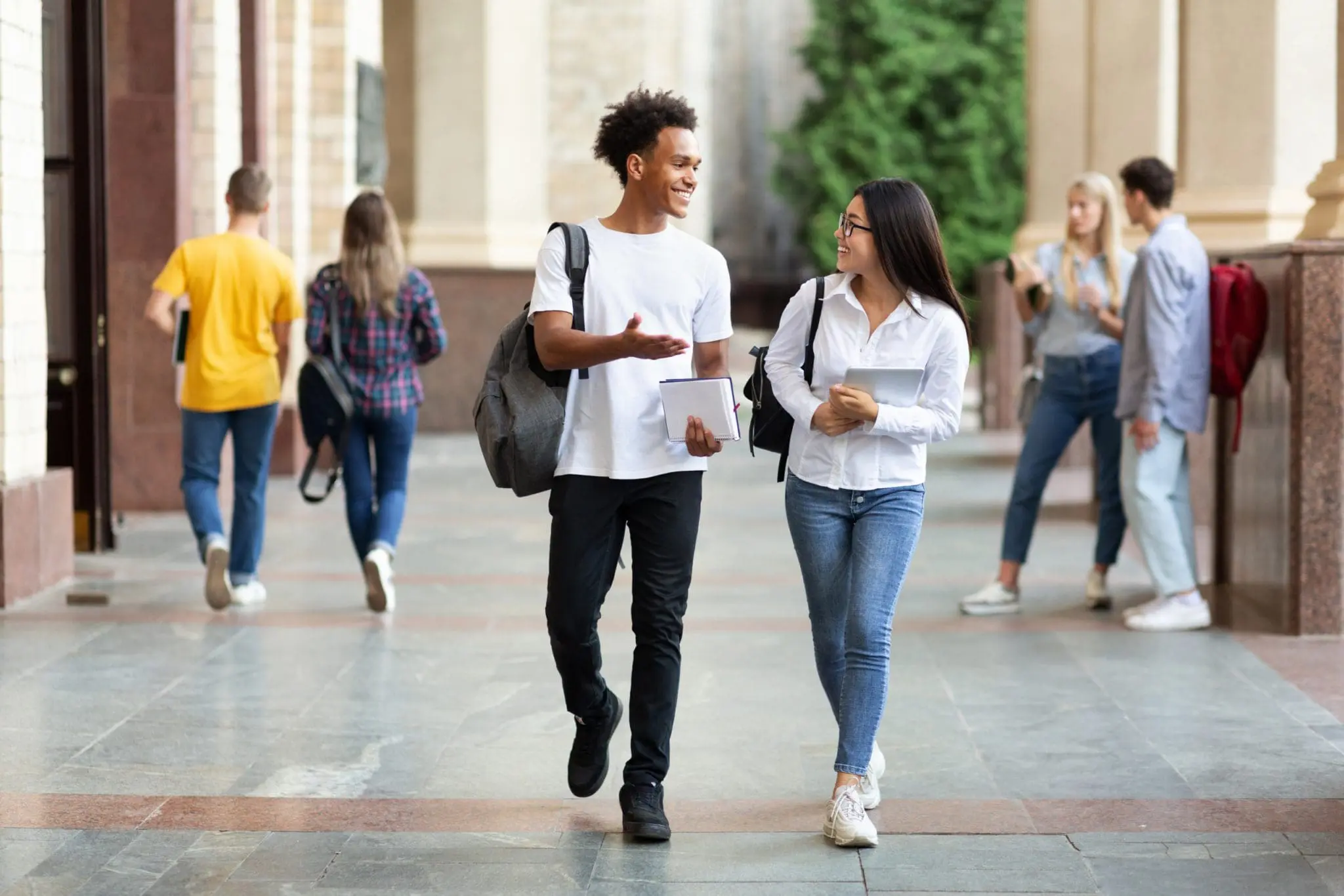 Two smiling students walking on campus and carrying books.
