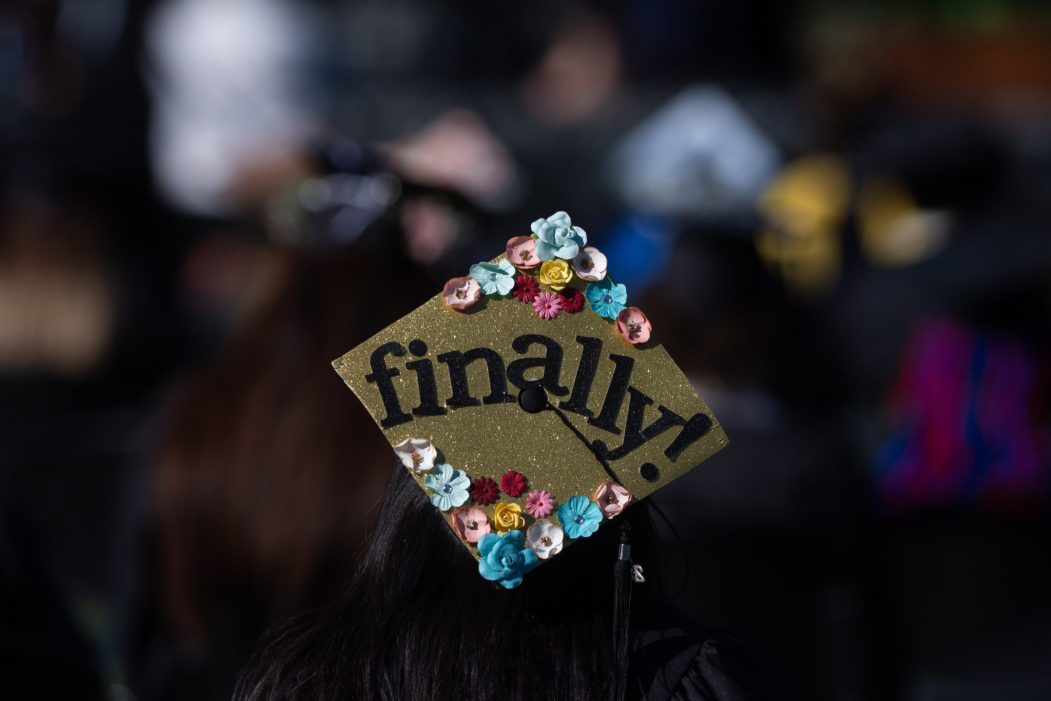 Students at a graduation ceremony. The focus is on a single graduation cap embellished with fake flowers, glitter, and the word Finally.