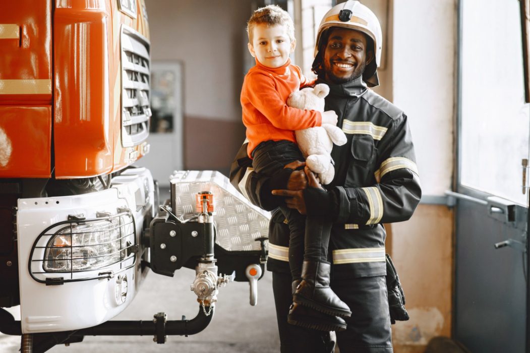Firefighter standing in front of a fire engine, holding a little boy, both of them smiling.